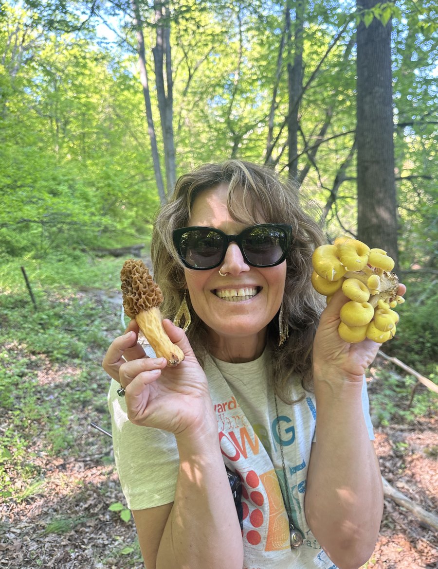 Laura deNey smiling in a sunlit forest, holding up a freshly foraged morel in one hand and a cluster of golden oyster mushrooms in the other.